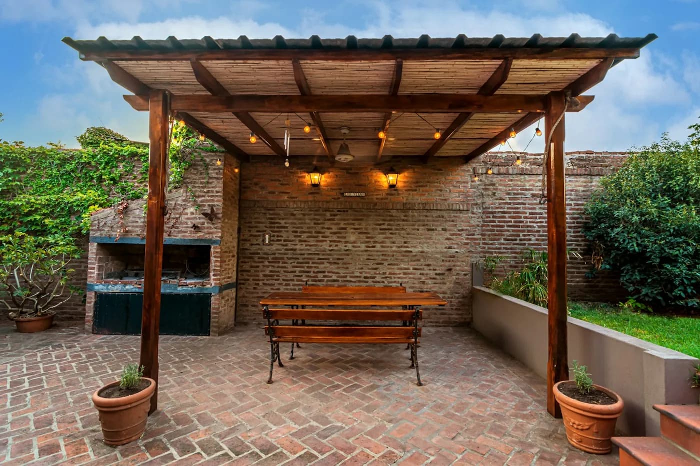 Espaço gourmet com churrasqueira de tijolos, pergolado de madeira com varal de luzes e mesa comprida com bancos